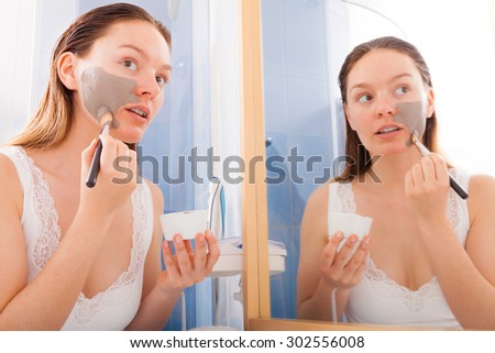 Beauty skin care cosmetics and health concept. Young woman holding brush and bowl with facial clay mask applying mud to her face in bathroom