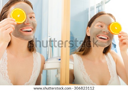Beauty skin care cosmetics and health concept. Young woman with facial clay mask holding orange fruit slice covering eyes in bathroom