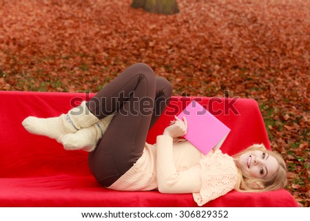 Happy smiling woman relaxing in fall park with book. Young blonde girl resting laying on bench. Autumn lifestyle fun.