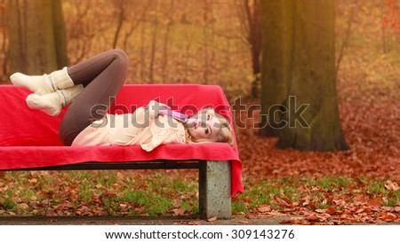 Happy smiling woman relaxing in fall park with book. Young blonde girl resting laying on bench. Autumn lifestyle fun.