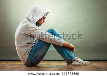 Young people and emotions concept. Sad hooded man teen boy with headphones sitting on floor grunge background