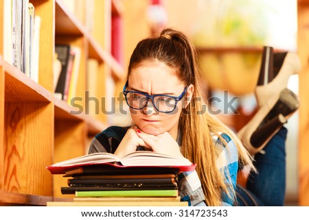 Education school concept. Clever female student girl in blue glasses lying on floor in college library with stack books reading. Indoor