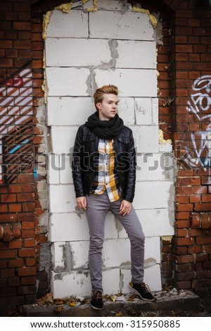 Full length of handsome trendy man outdoor in city setting, male model wearing black jacket scarf and checked shirt against brick wall
