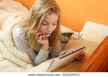 Lazy young woman using computer tablet browsing internet. Girl laying in bed under blanket and holding ebook. Technology leisure.