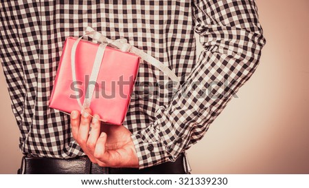 Man hiding pink gift box with white ribbon behind back. Closeup of male hand holding christmas present. Guy wearing flannel shirt. Birthday, holiday surprise. Instagram filtered.