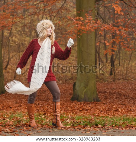 Fashion woman with flying scarf in windy fall autumn park forest against blowing wind. Young girl posing in fur cap and sweater having fun outdoor.