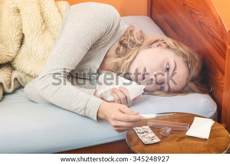 Sick woman laying in bed under wool blanket sneezing in tissue holding thermometer. Ill girl caught cold flu. Pills and tablets on table.