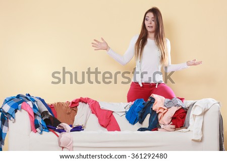 Helpless woman standing behind on sofa couch in messy living room shrugging. Young girl surrounded by many stack of clothes. Disorder and mess at home.