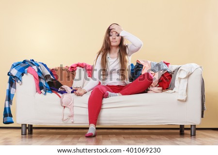 Desperate helpless woman sitting on sofa couch in messy living room with hand on head. Young girl surrounded by many stack of clothes. Disorder and mess at home.