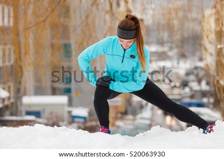 Girl wearing sportswear and doing stretching exercises on snow with urban background. Winter sports, outdoor fitness, fashion, workout, health concept.