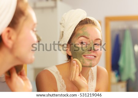 Woman removing facial dried clay mud mask with sponge in bathroom in front of mirror. Skin care. Girl taking care of her complexion. Beauty spa treatment.