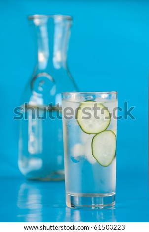 Glass of ice water with sliced cucumbers and a carafe on teal blue