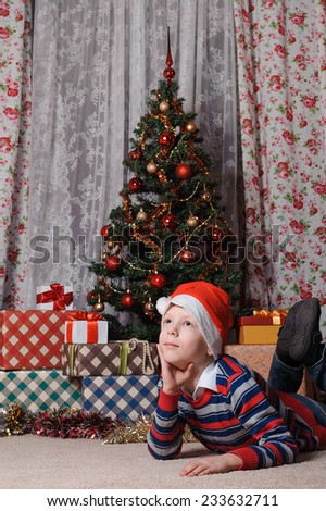 adorable boy in Santa hat dreaming about the  New Year\'s holidays near christmas tree