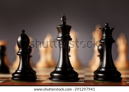 Behind the scenes - black chess king, queen and bishop in focus standing on a wooden chessboard facing the white chess pieces before the game