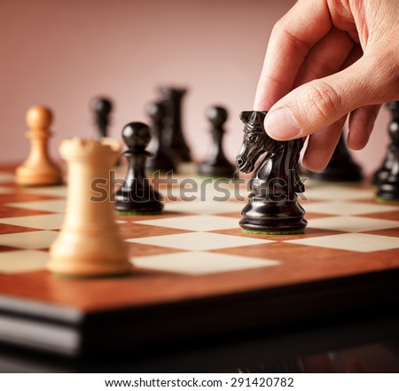 Male hand moving the black chess knight in focus in the middle of a chess game with blurred white rook in the foreground