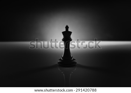 Solitary black chess king carved in genuine ebony wood in focus standing on a glossy table in the dark