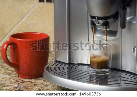 Espresso machine pouring shot into shot glass with red mug standing by.
