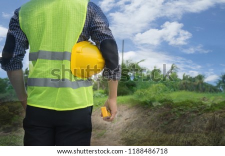 Back view of engineer, architect, supervisor worker, wearing green reflective vest, holding yellow safety helmet, water level gauge at construction site, empty land. Real estate, Home building concept