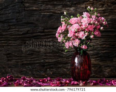 Still life visual art of pink roses arrangement in red glass vase on wooden slap with dry flowers falling round and old wooden back drop