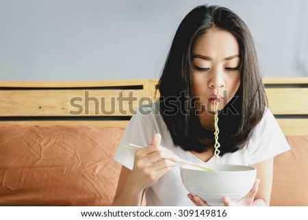Asian girl eats instant noodle on the bed and noodle still in her mouth.