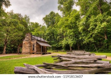 An old log and stone cabin in the green woods with an old wooden fence in the foreground and a grey sky