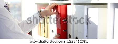 Male arms pick red file folder from office book shelf closeup. Store pile of project documentation concept