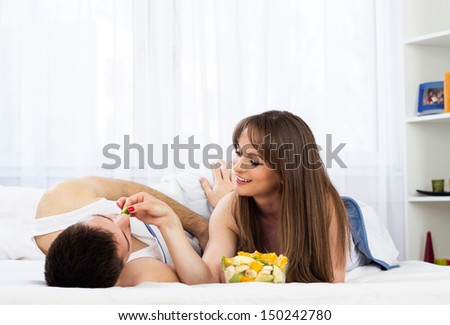 Young couple eating fruit salad. Young female feeding his partner with fruit salad in bedroom