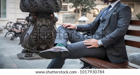 Suited man sitting on bench outdoors with his leg crossed and with his hands on his legs