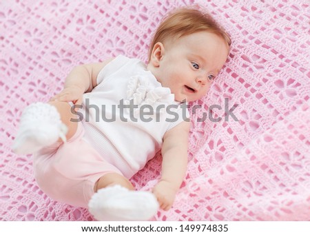 The baby lies on a pink towel. She\'s in pink shorts and a white blouse.