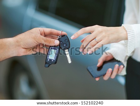 Male Hand Giving Car Key To Female Hand. She Is Holding A Cell Phone. In The Background, A Fragment Of The Car.