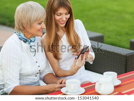 Adult mother and daughter. The daughter on the phone shows a photo of the fiance. They drink coffee and tea.