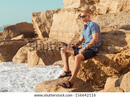 Fashionable young muscular guy resting on a rocky beach. He is sitting on a rock, dressed in flowing blue shirt and sunglasses.