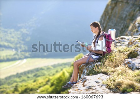 Young, beautiful girl with a backpack on her back, studying a map while standing on the plateau. In the background, green meadows and majestic mountains.