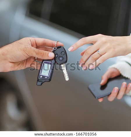 Male hand giving car key to female hand. She is holding a cell phone. In the background, a fragment of the car.
