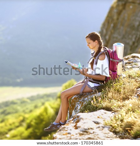 Young, Beautiful Girl With A Backpack On Her Back, Studying A Map While Standing On The Plateau. In The Background, Green Meadows And Majestic Mountains.