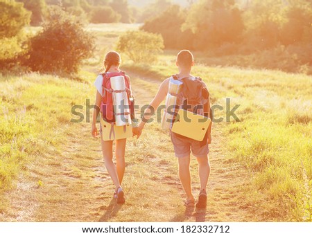Two tourists with backpacks on the plateau. Young couple go on the road hand in hand. The sun\'s rays setting sun illuminates them. In the background, mountains covered with forests.