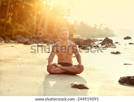 Fit young man practices sun salutation yoga on the beach at sunset