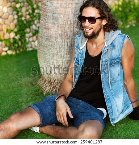 fashion man in sunglasses and jeans clothes sits on grass near palms and smiling Vogue