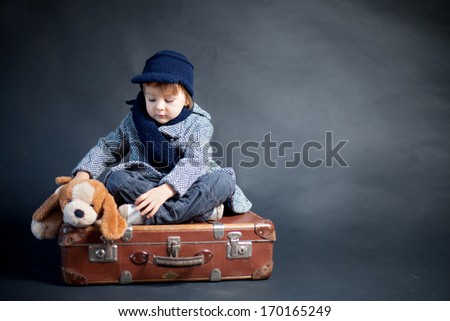 Little boy with coat and hat, sitting on a suitcase with his teddy dog friend