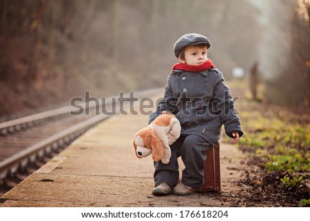 Portrait of a little boy with s suitcase and a teddy dog, waiting on a railway station