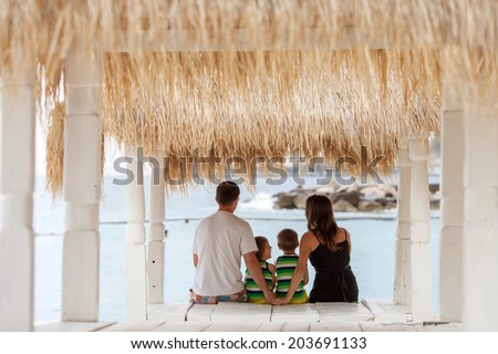 Back view of a happy family, sitting on the beach, watching the sea