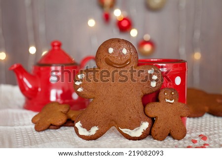 Smiling gingerbread man and a smaller one next to him standing in front of a mug. Teapot and more cookies on the background. Lights and christmas decoration in the background. Red and brown colors