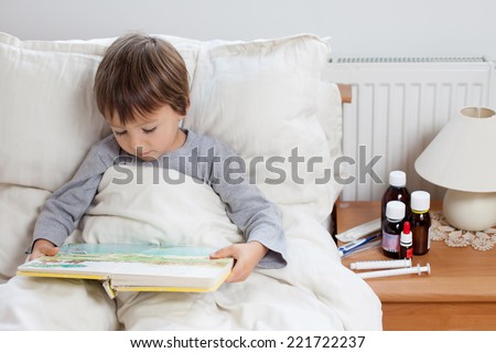 Sick child boy lying in bed with a fever, resting reading a book