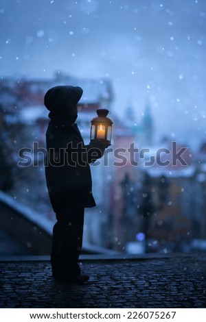 Silhouette of boy, standing on stairs, holding lantern, view of Prague behind him, snowy evening