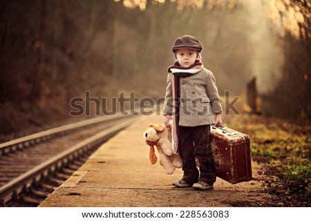Adorable boy on a railway station, waiting for the train with suitcase and teddy bear