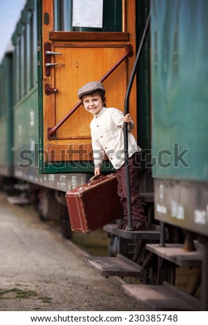 Boy, dressed in vintage shirt and hat, with suitcase, on a railway station, standing on a steam train stairs, looking out