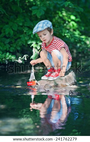 Cute little boy playing on a pond with a toy boat in hands in a warm sunny summer day.