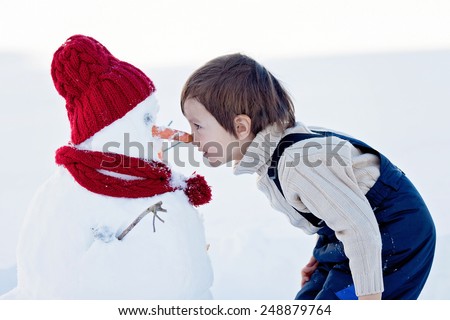 Happy beautiful child building snowman in garden, winter time, nose to nose with the snowman