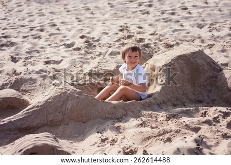 Adorable kid, playing on the beach, sitting in a ferrari car, made of sand, smiling at the camera, pretending to drive