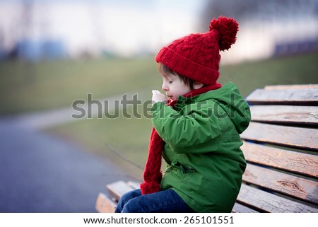 Little boy sneezing and blowing his nose outdoor on a sunny winter day sitting on a bench Little boy sneezing and blowing his nose outdoor on a sunny winter day sitting on a bench